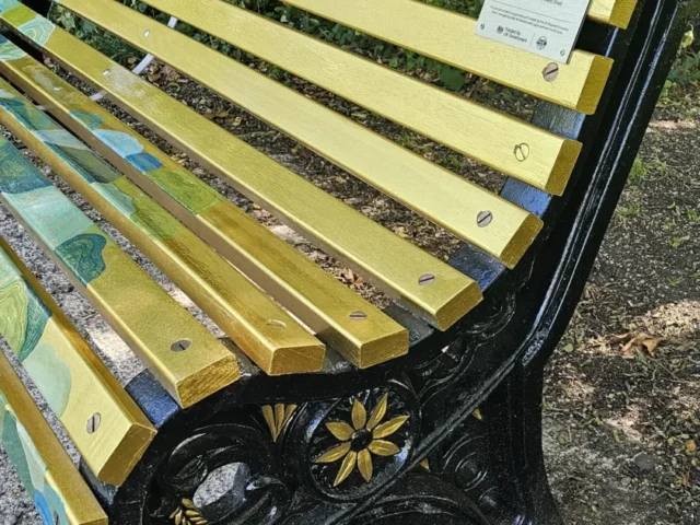 Close view of the golden painted timber slats, black metal frame and artist plaque on Jade Dawn’s River Lea bench.