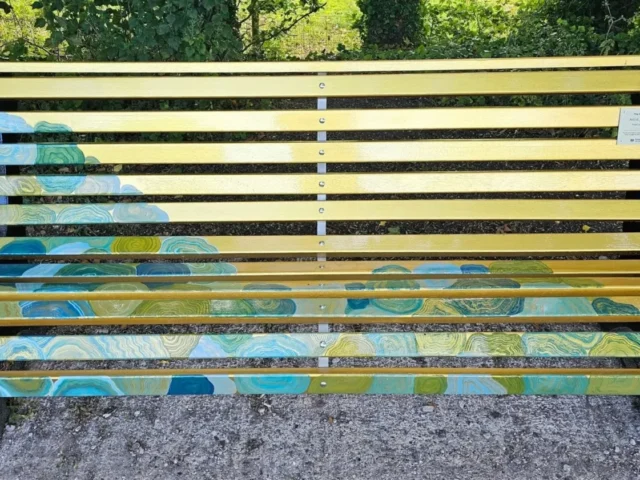 Full front view of Jade Dawn’s completed golden bench with blue and green river-inspired artwork across the slats.