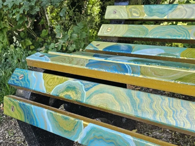 Close-up of blue, green and gold marbled artwork across the timber slats of the River Lea Art Trail bench.